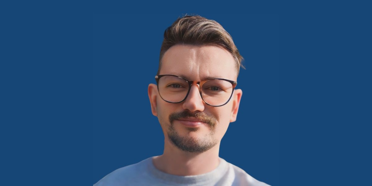 A young white man with a blond quiff, a beard and moustache wearing glasses on a blue background